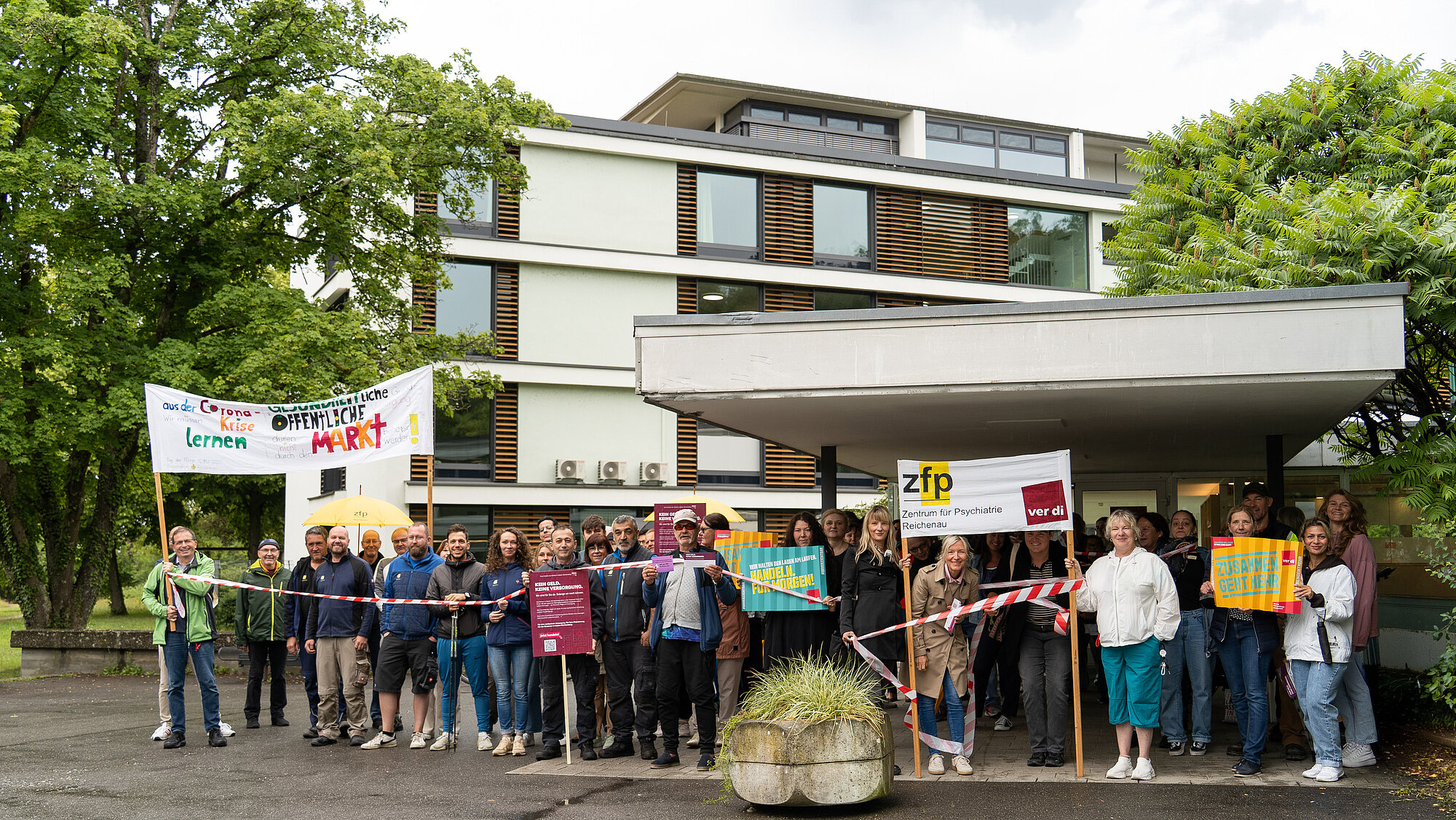Eine große Gruppe von Menschen steht mit Plakaten und Absperrband vor einem Klinikgebäude.