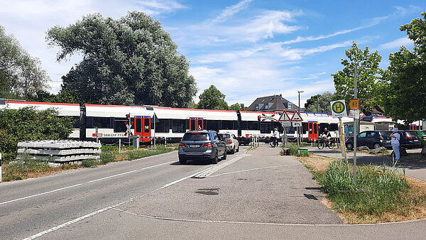 Autos, Radfahrer und Fußgänger stehen vor einem geschlossenen Bahnübergang und ein Zug fährt vorbei.