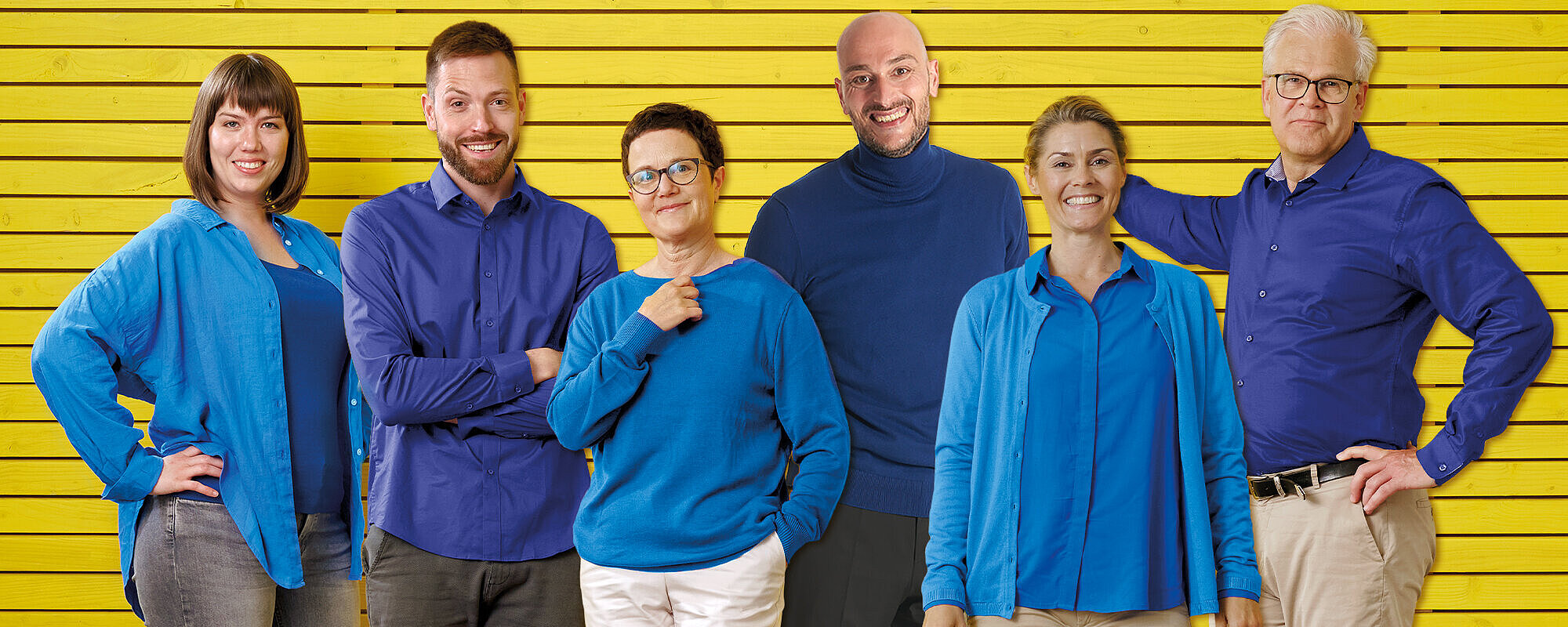 Eine Gruppe von Frauen und Männern in hell- und dunkelblauer Kleidung steht gemeinsamer vor einem gelben Hintergrund.