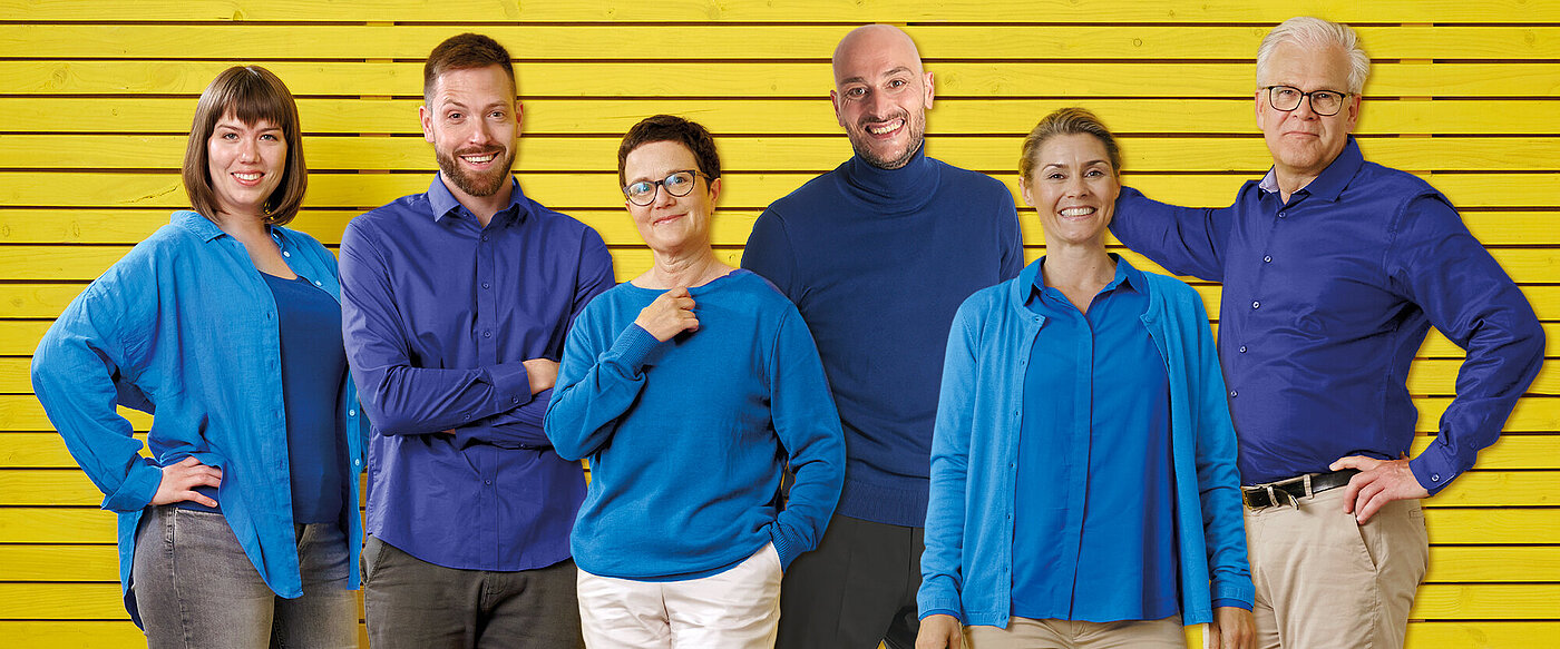 Eine Gruppe von Frauen und Männern in hell- und dunkelblauer Kleidung steht gemeinsamer vor einem gelben Hintergrund.