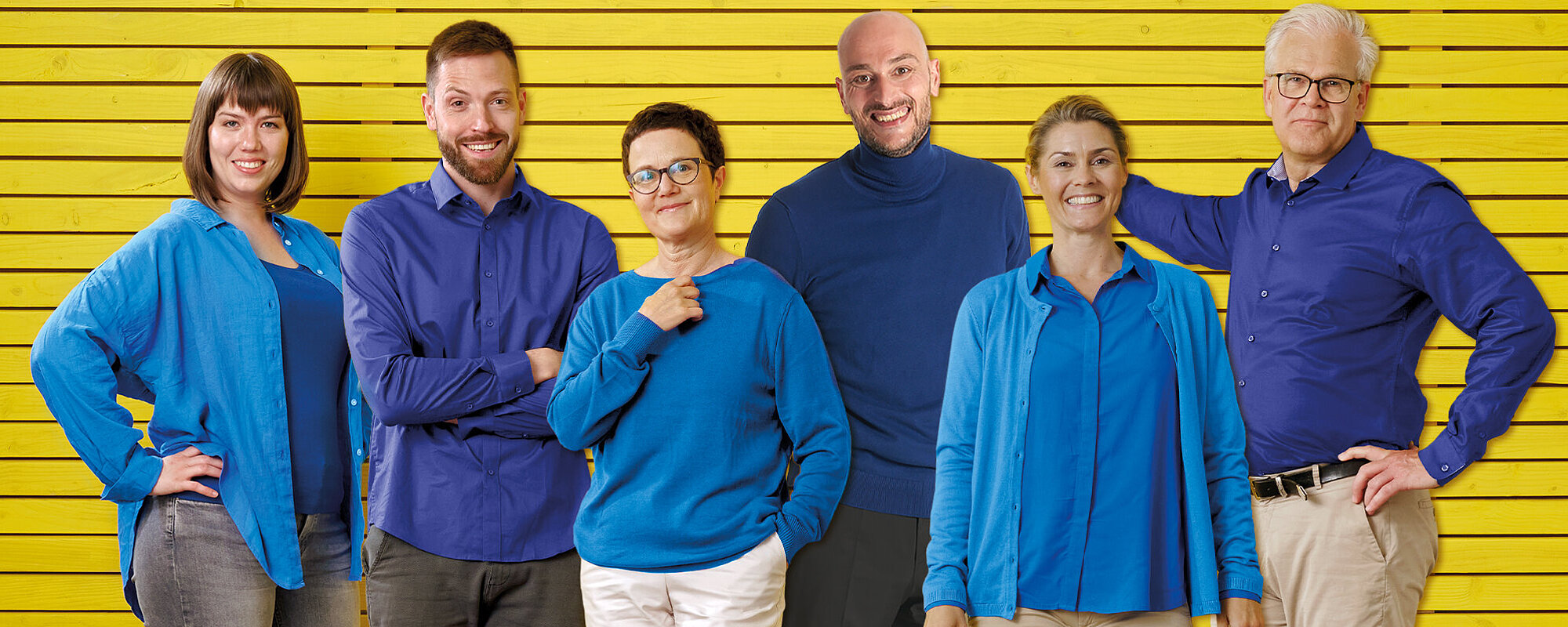 Eine Gruppe von Frauen und Männern in hell- und dunkelblauer Kleidung steht gemeinsamer vor einem gelben Hintergrund.