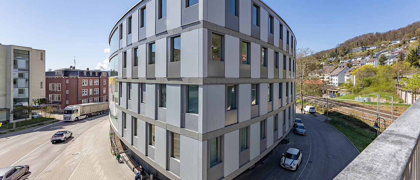 Vierstöckiges, ovales Gebäude aus Glas und Beton im Herzen einer Stadt und an einer Kreuzung gelegen.