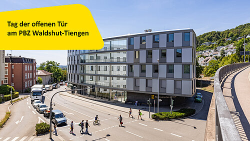 Das Bild zeigt ein großes mehrstöckiges Gebäude an einer Straßenkreuzung mit Autos, Menschen und einem gelben Einklinker mit Text.