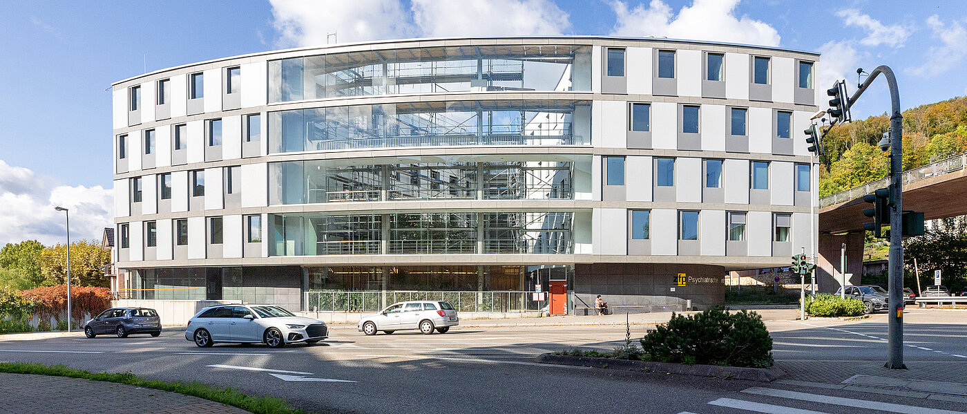 Vierstöckiges, ovales Gebäude aus Glas und Beton an einer befahrenen Kreuzung.
