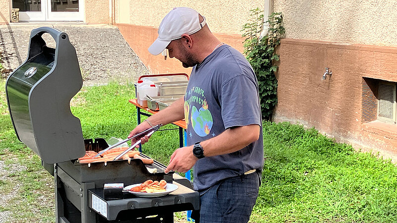 Ein Mann mit Basecap steht an einem Gasgrill und grillt verschiedene Würstchen.