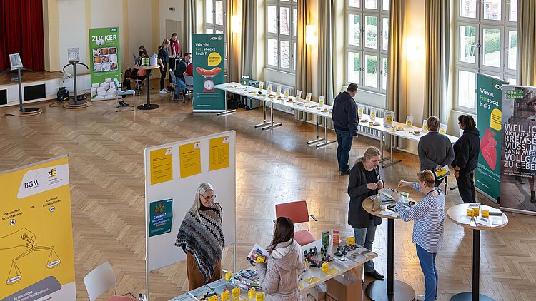 In einem Saal mit großen Fenstern stehen verschiedene Stände mit Tischen und Roll-ups, die von Menschen besucht werden.