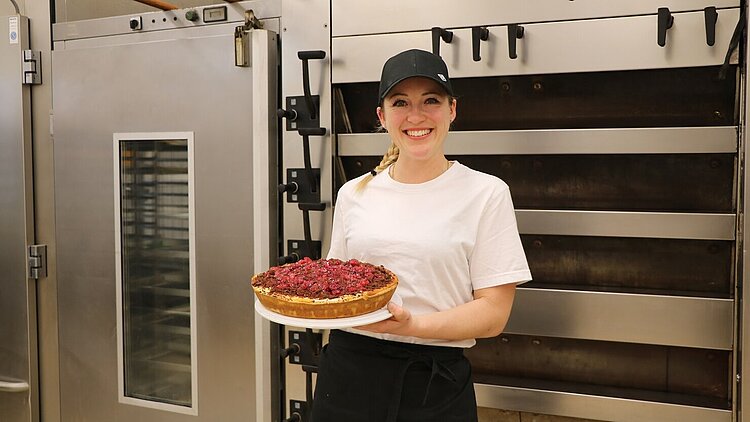 Eine Frau mit weißem T-Shirt und schwarzer Mütze und Schürze steht in einer Backstube und hält einen Kuchen mit roten Früchten.
