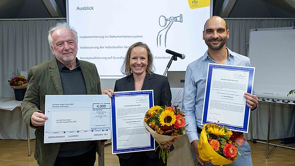 Drei Menschen halten Urkunden und einen Scheck in der Hand und lächeln in die Kamera.