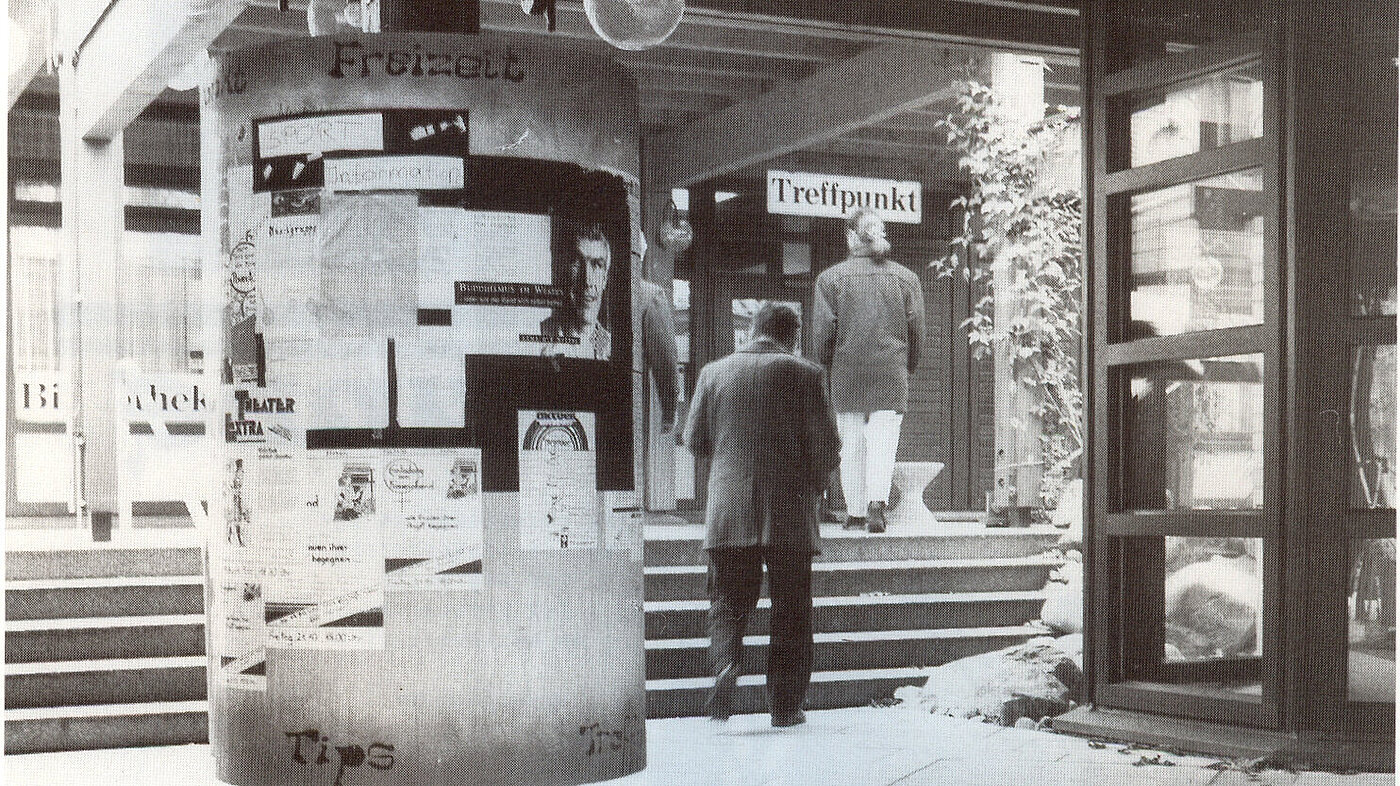 Schwarz-weiß Foto einer Litfasäule mit verschiedenen Plakaten und Menschen im Hintergrund, die eine Treppe hochgehen. 