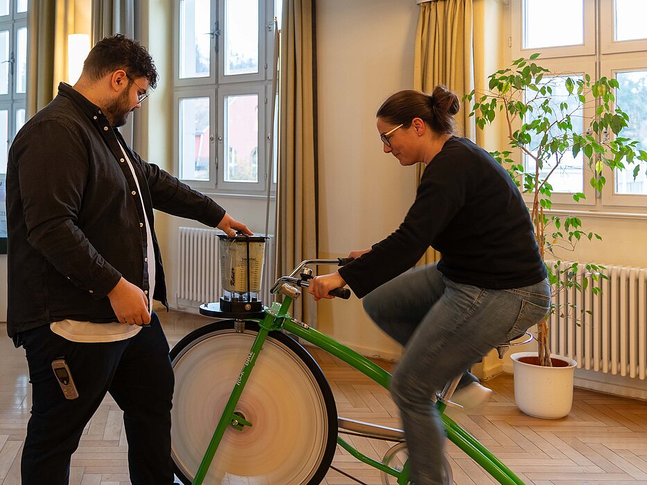 Eine Frau sitzt auf einem grünen Fahrrad und radelt, um mit ihrer Muskelkraft die Zutaten in einem Mixer zu zuerkleinern.