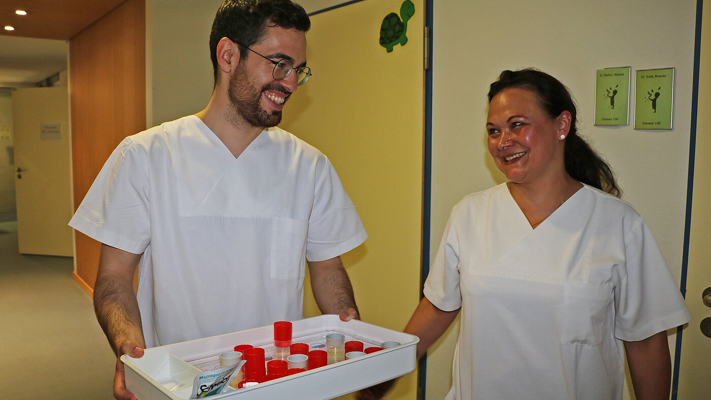Ein Mann und eine Frau in weißer Pflegekleidung lachen einander an. Er hält ein Tablett mit roten und transparenten Bechern mit Medikamenten in der Hand.