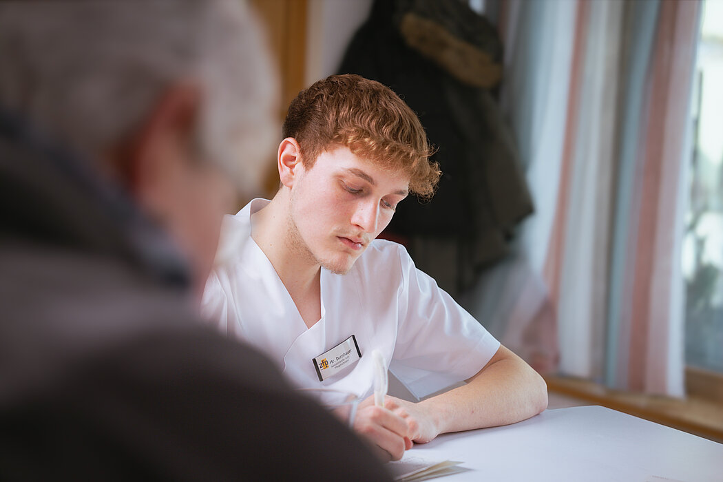 Ein junger Mann in Pflegekleidung sitzt konzentriert am Tisch und macht Notizen.
