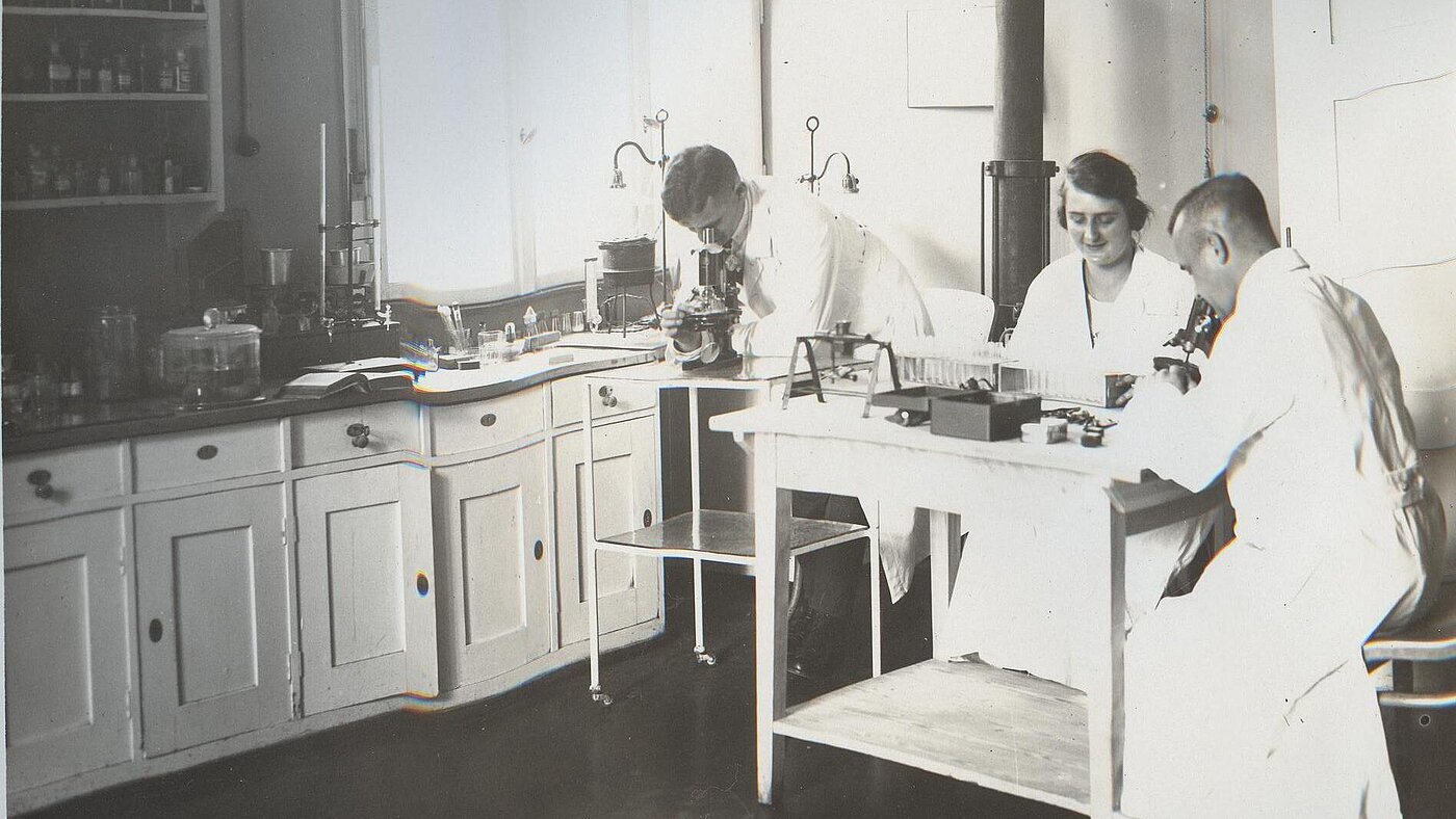 Schwarz-weiß Foto, das zwei Männer und eine Frau in einem alten, historischen Labor beim Arbeiten zeigt.