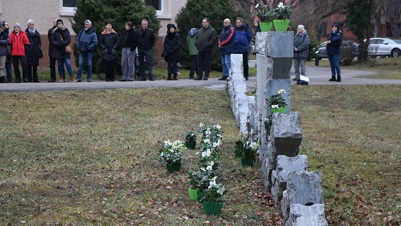 Im Vordergrund ist seitlich das Mahnmal mit den Blumen zu sehen, im Hintergrund stehen Teilnehmende.