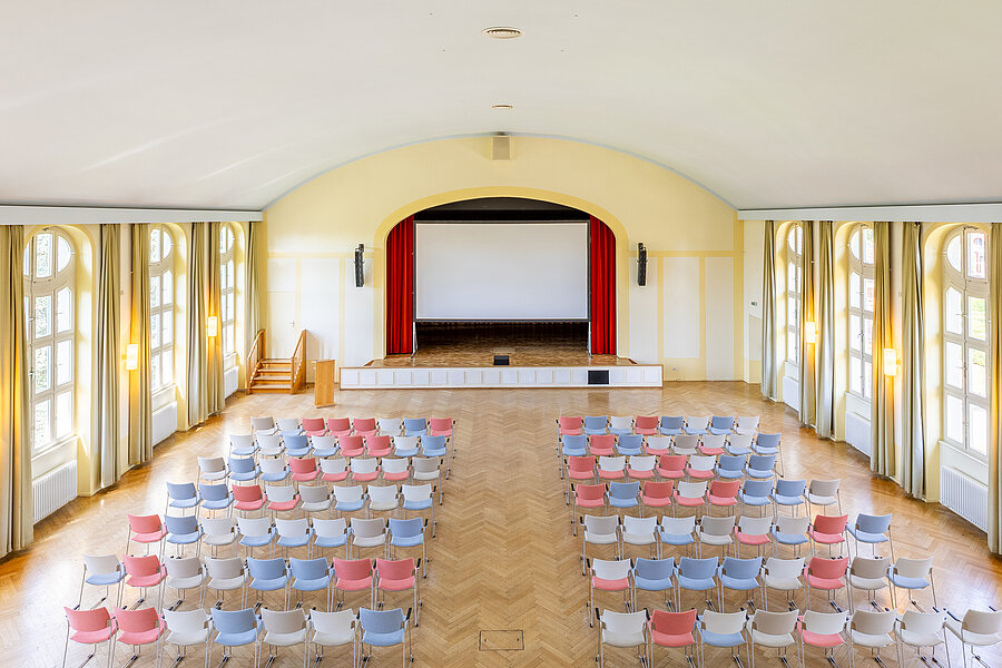 Blick von einer Empore in einen bunt bestuhlten Festsaal mit Bühne.