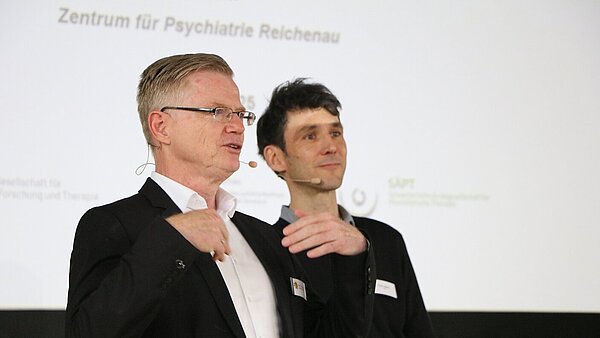 Zwei Männer, beide mit einem Mikrofon, stehen auf einer Bühne und im Hintergrund ist eine Präsentation zu sehen.