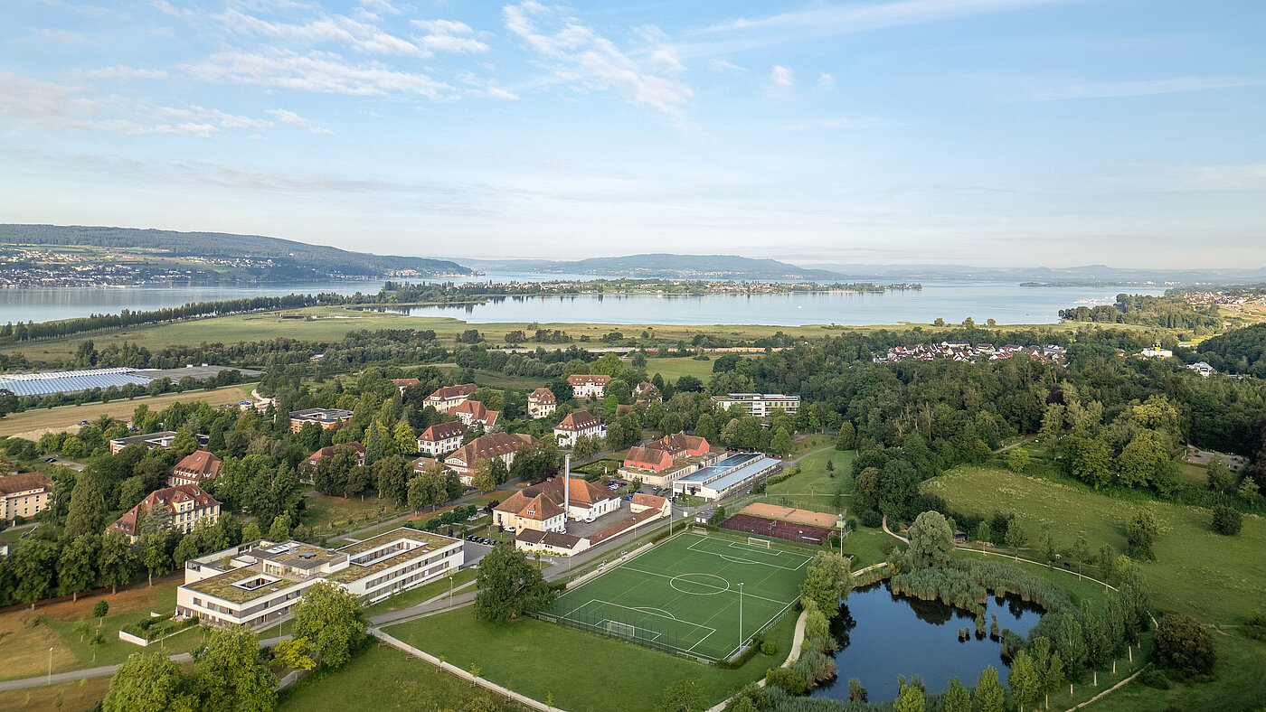 Luftbildaufnahme des ZfP Reichenau mit den einzelnen Häusern, dem Sportplatz sowie dem Teich und im Hintergrund dem Bodensee und den Bergen.