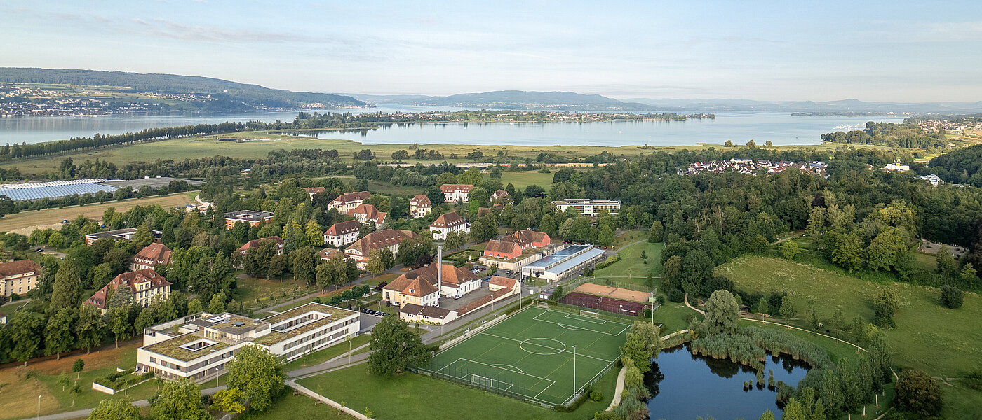 Luftbildaufnahme des ZfP Reichenau mit den einzelnen Häusern, dem Sportplatz sowie dem Teich und im Hintergrund dem Bodensee und den Bergen.