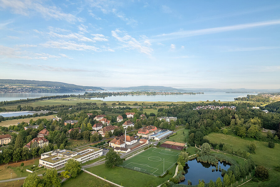 Luftbildaufnahme des ZfP Reichenau mit den einzelnen Häusern, dem Sportplatz sowie dem Teich und im Hintergrund dem Bodensee und den Bergen.