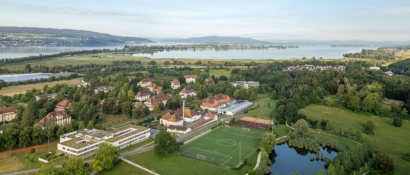 Luftbildaufnahme des ZfP Reichenau mit den einzelnen Häusern, dem Sportplatz sowie dem Teich und im Hintergrund dem Bodensee und den Bergen.