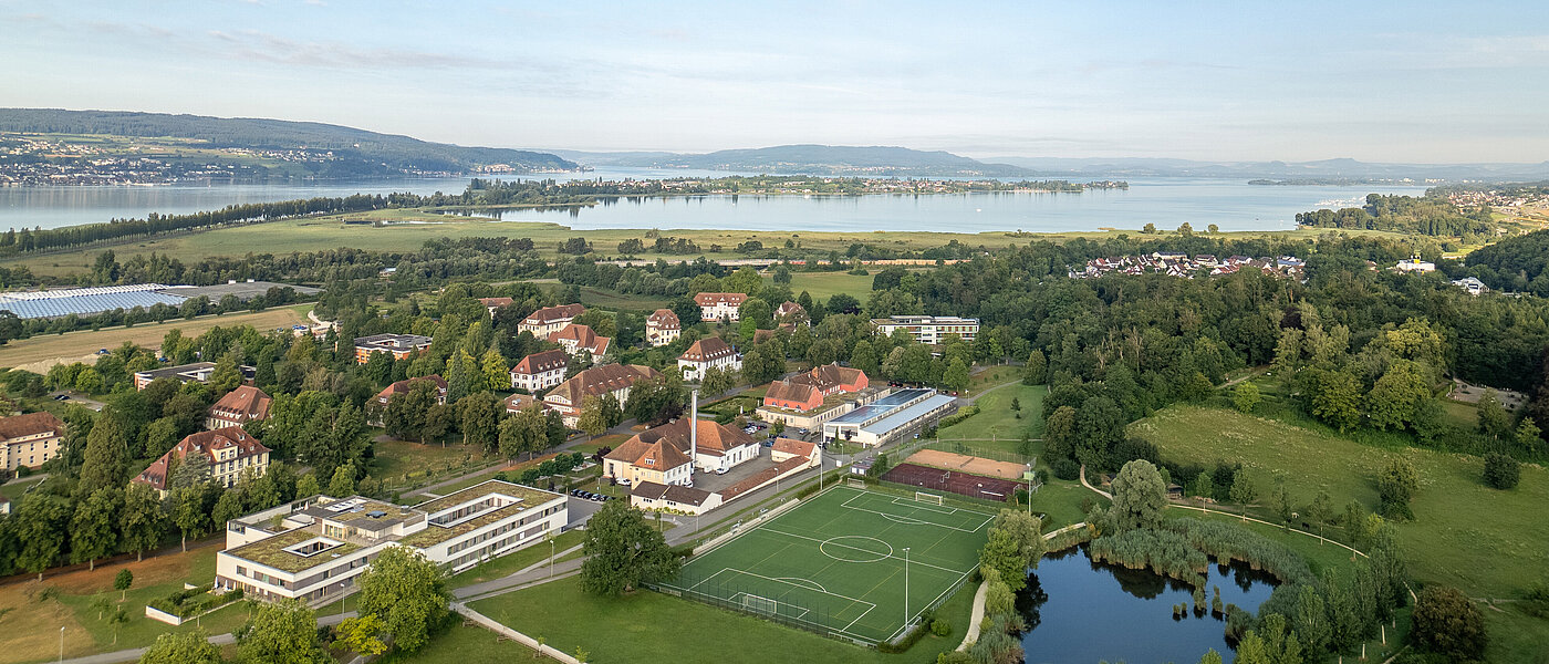 Luftbildaufnahme des ZfP Reichenau mit den einzelnen Häusern, dem Sportplatz sowie dem Teich und im Hintergrund dem Bodensee und den Bergen.