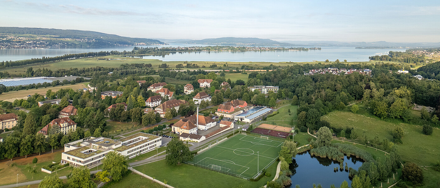 Luftbildaufnahme des ZfP Reichenau mit den einzelnen Häusern, dem Sportplatz sowie dem Teich und im Hintergrund dem Bodensee und den Bergen.