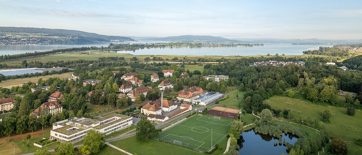 Luftbildaufnahme des ZfP Reichenau mit den einzelnen Häusern, dem Sportplatz sowie dem Teich und im Hintergrund dem Bodensee und den Bergen.