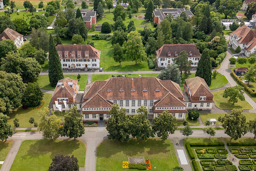 Luftbildaufnahme von einem weitläufigen Gelände mit verschiedenen Häusern, Parkplätzen und dem Bodensee im Hintergrund.