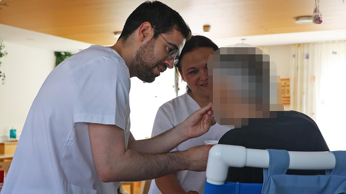 Ein Mann und eine Frau stehen vor einer sitzenden Person. Der Mann füttert die Person.