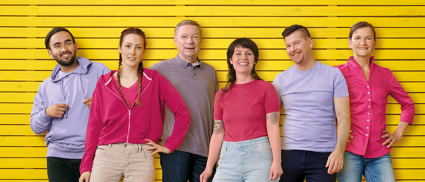 Eine gruppe von Männern und Frauen in flieder- und pinkfarbener Kleidung vor einer gelben Wand. 