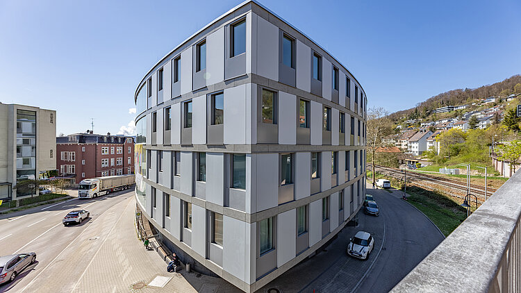 Vierstöckiges, ovales Gebäude aus Glas und Beton im Herzen einer Stadt und an einer Kreuzung gelegen.