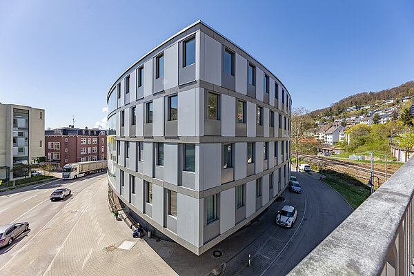 Vierstöckiges, ovales Gebäude aus Glas und Beton im Herzen einer Stadt und an einer Kreuzung gelegen.