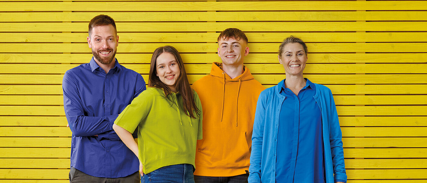 Eine Gruppe, bestehend aus einer Frau, einem Mann, einem Mädchen und einem Jungen, stehen vor einer gelben Holzwand.