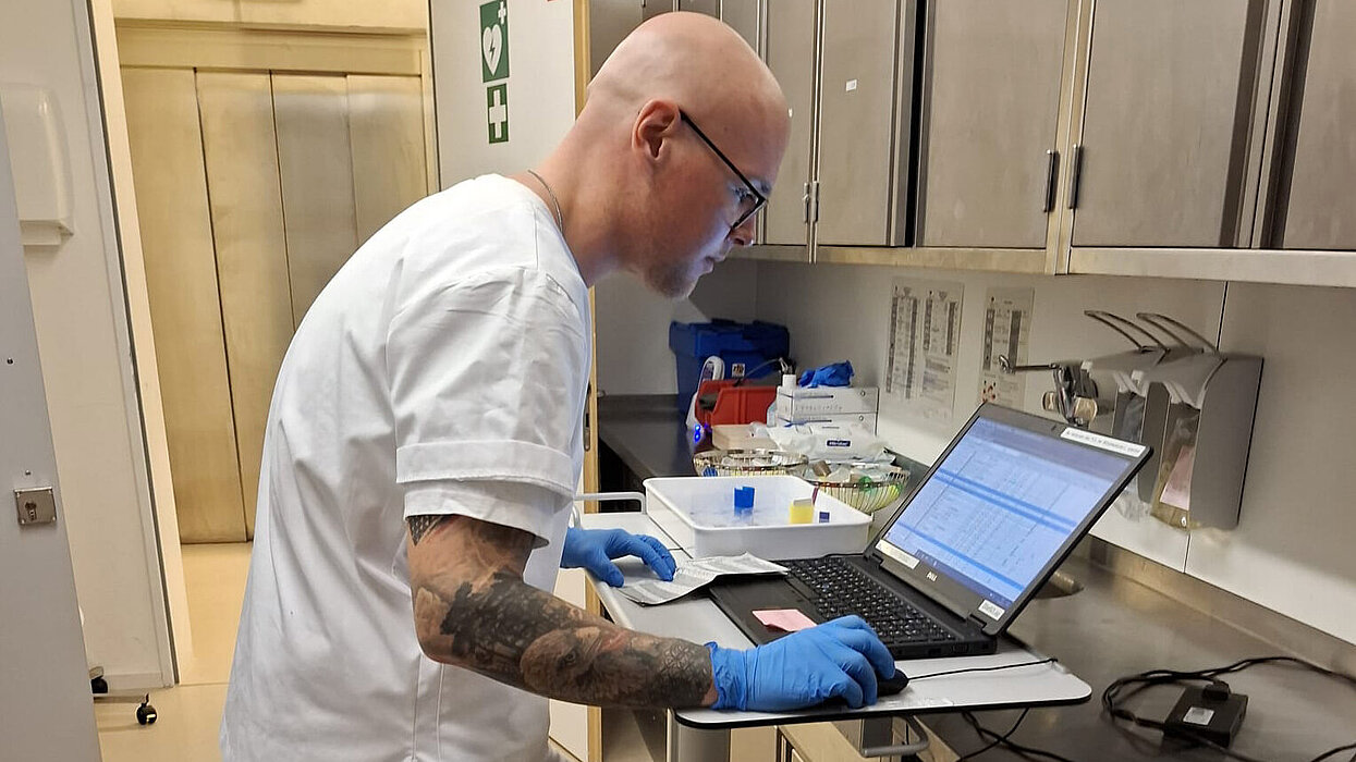Ein junger Mann mit tätowiertem Arm und in Pflegekleidung steht an einem Tisch und blickt in ein Laptop.