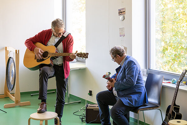 Zwei Männer, einer stehend und einer sitzend, spielen Gitarre.