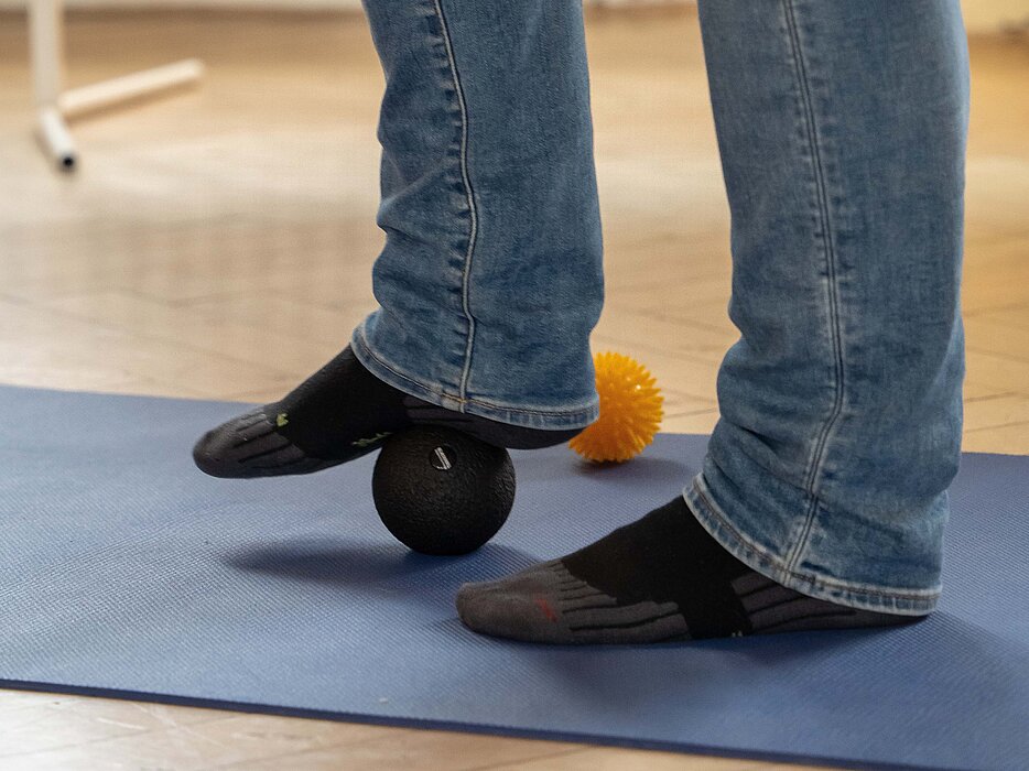 Eine Person in Jeanshosen und Strümpfen rollt den Fuß über einen Massageball.