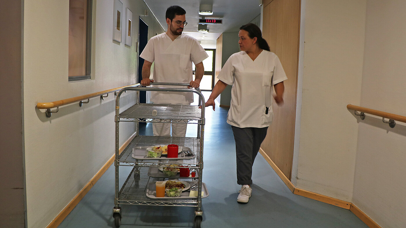 Ein Mann schiebt einen Wagen mit portionierten Tellergerichten durch einen Flur. Neben ihm läuft eine Frau.