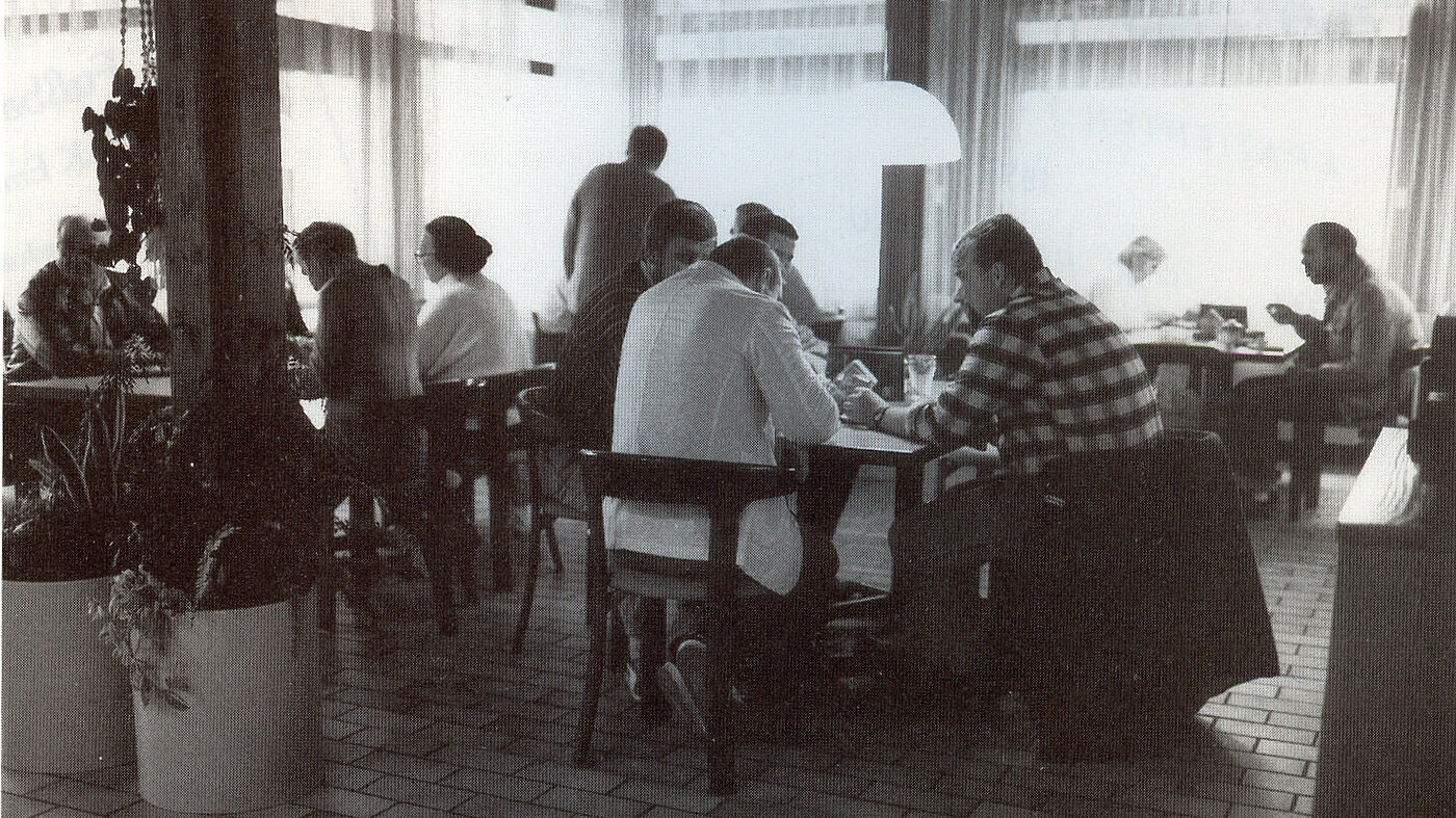 Schwarz-weiß Foto von Menschen, die in verschiedenen Gruppen an Tischen sitzen und essen.