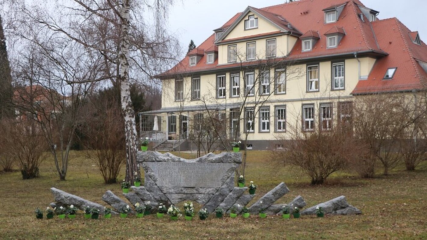 Chrstrosen stehen vor und auf dem Mahnmal, das aus einem zentralen Granitblock und seitlichen, schräg stehenden Stelen besteht.