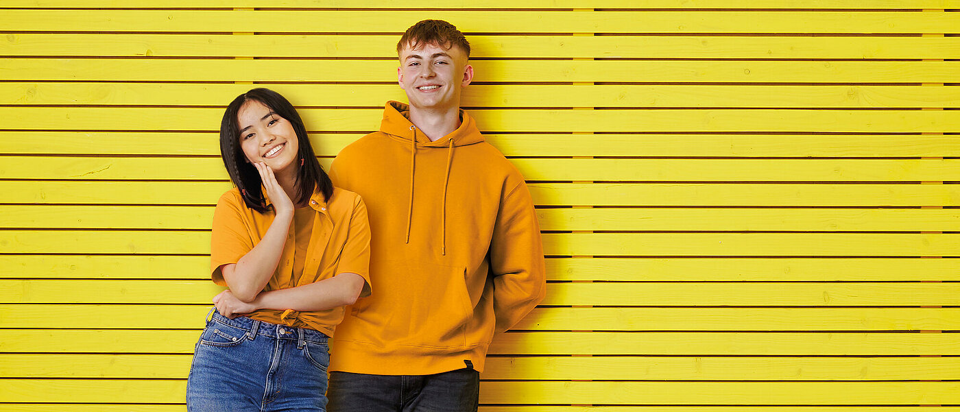 Ein Mädchen und ein junge in orangefarbener Kleidung stehen gemeinsam vor einer gelben Wand. 