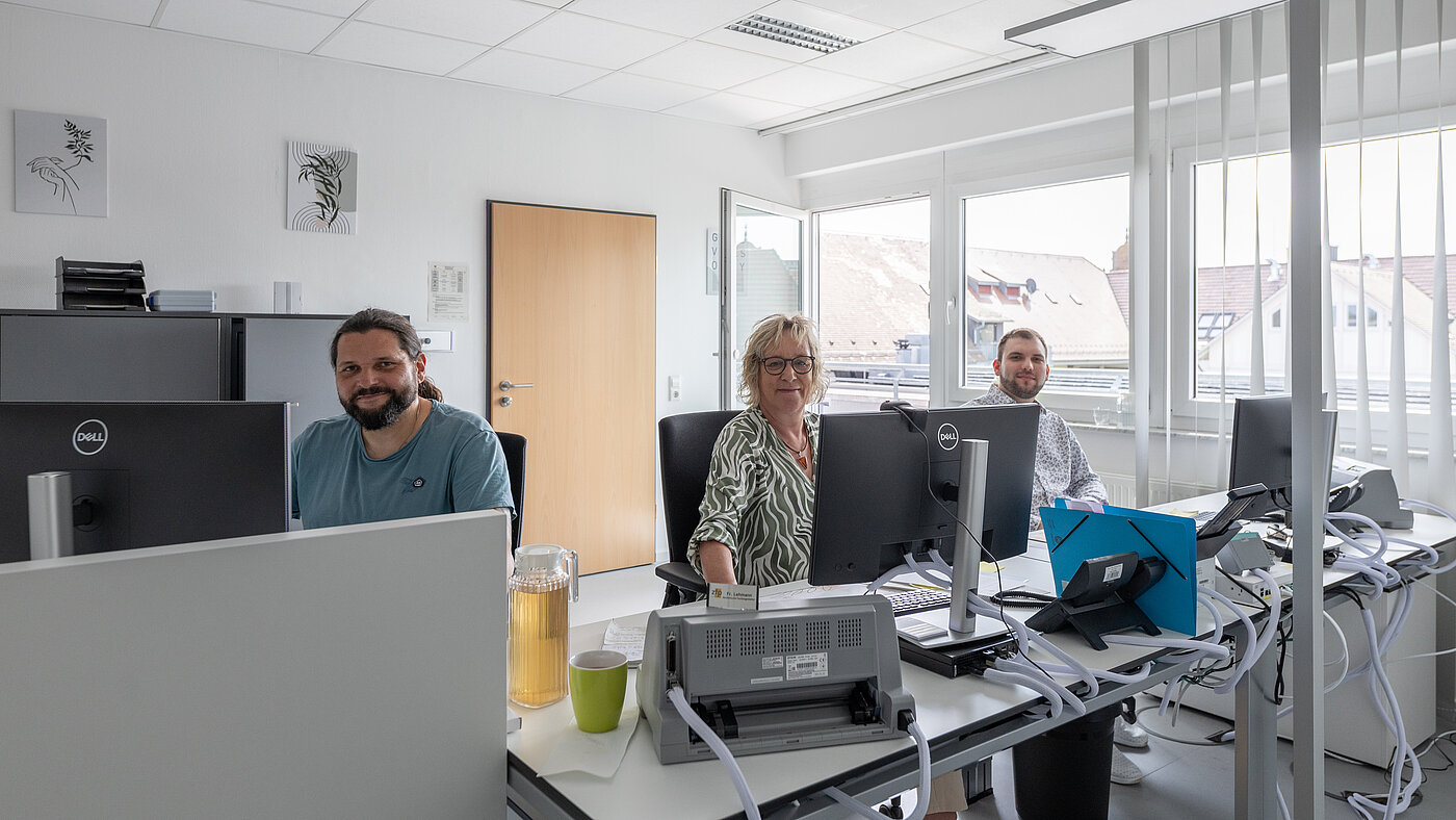 Zwei Männer und eine Frau sitzen in einem Büro hinter ihren Schreibtischen an Computern.