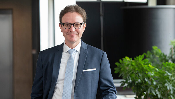 Ein lächelnder Mann in blauem Anzug und Brille lehnt an einem Glastisch.