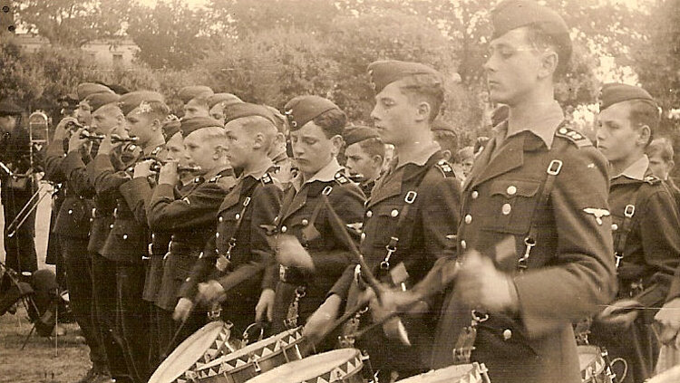 Schwarz-weiß Foto, das Jungs zeigt, die in Uniformen in einer Reihe stehen und und Trommeln schlagen.